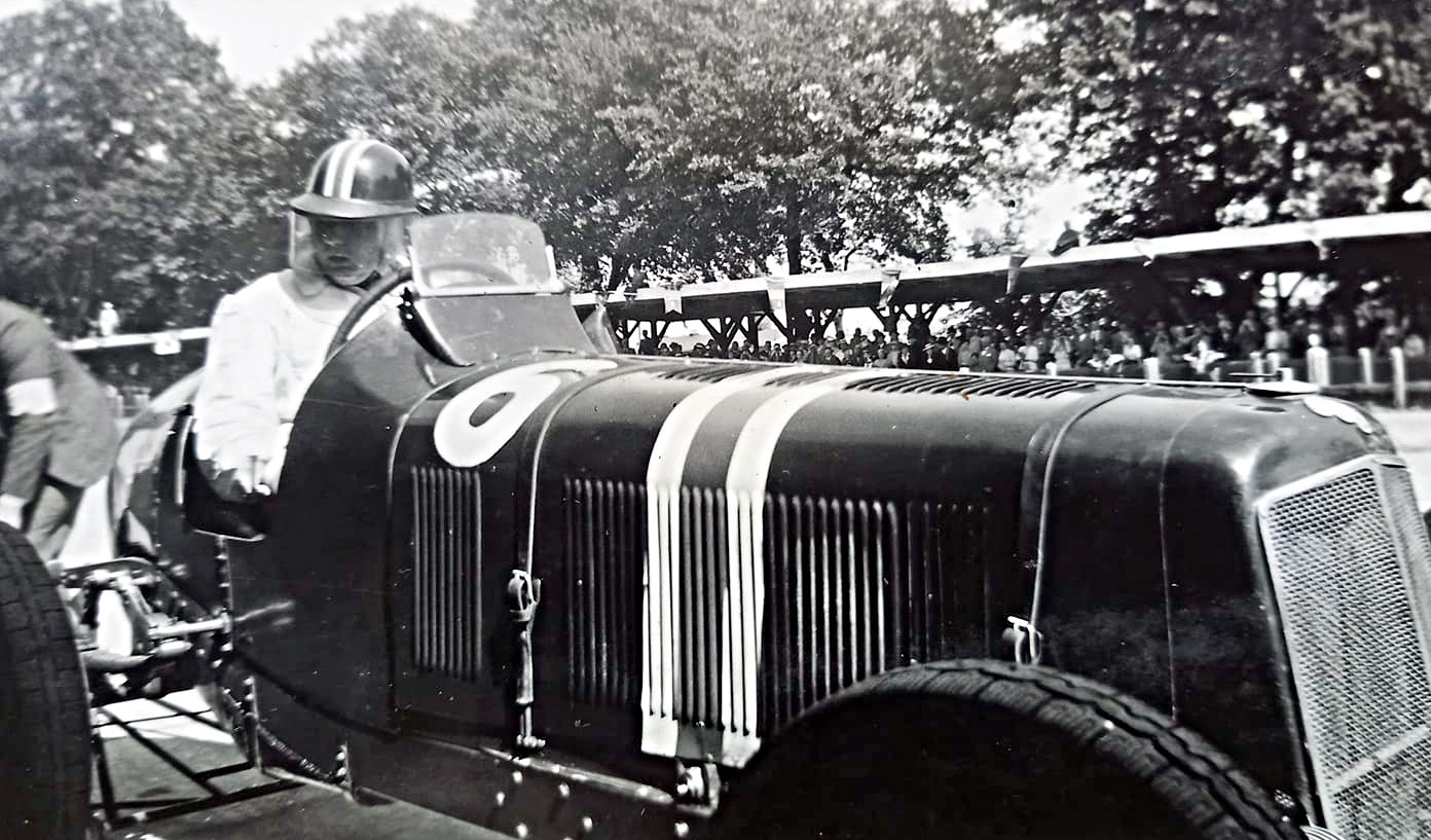 Black-and-white photograph of the dark ERA R1A with white stripes on its bonnet, its helmeted driver seated, in the Avus paddock with a grandstand behind