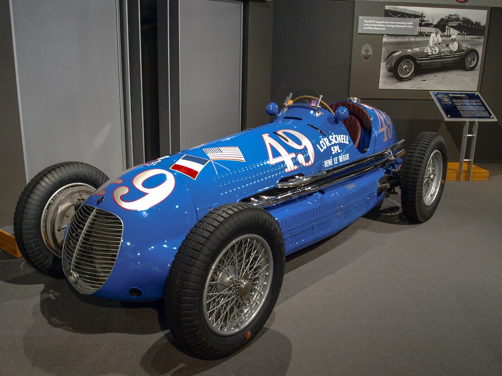A bright blue Maserati 8CTF numbered 49 with French and American flags on display in a museum, with a period photograph of the car on the wall behind