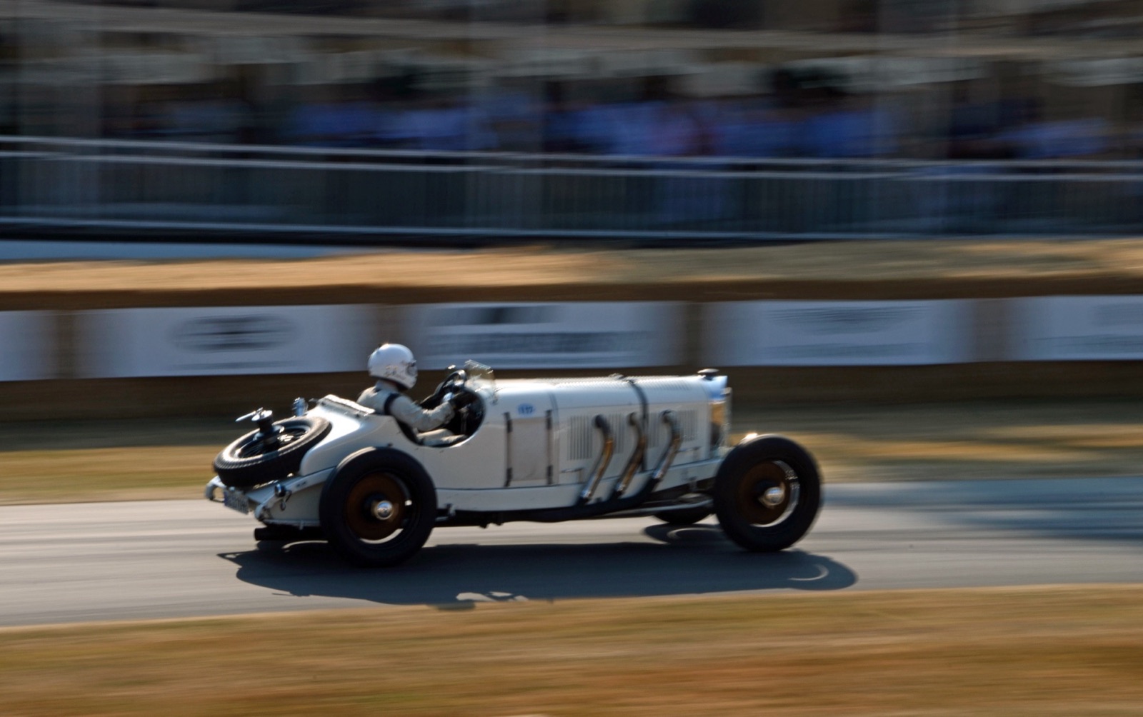 A white Mercedes-Benz SSK with a spare wheel on its tail and three external exhaust pipes on the side of its long bonnet, driven fast past a fence