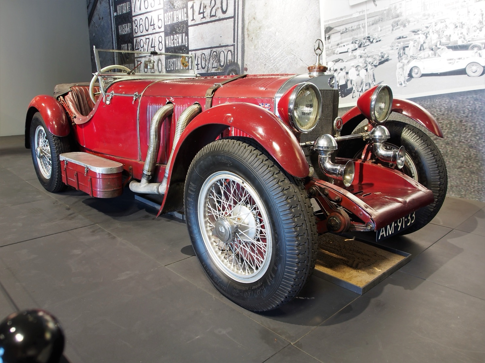 A red Mercedes-Benz SSK two-seater with worn paint, flexible exhaust pipes and wire wheels in a museum, in front of a wall printed with large factory record numbers