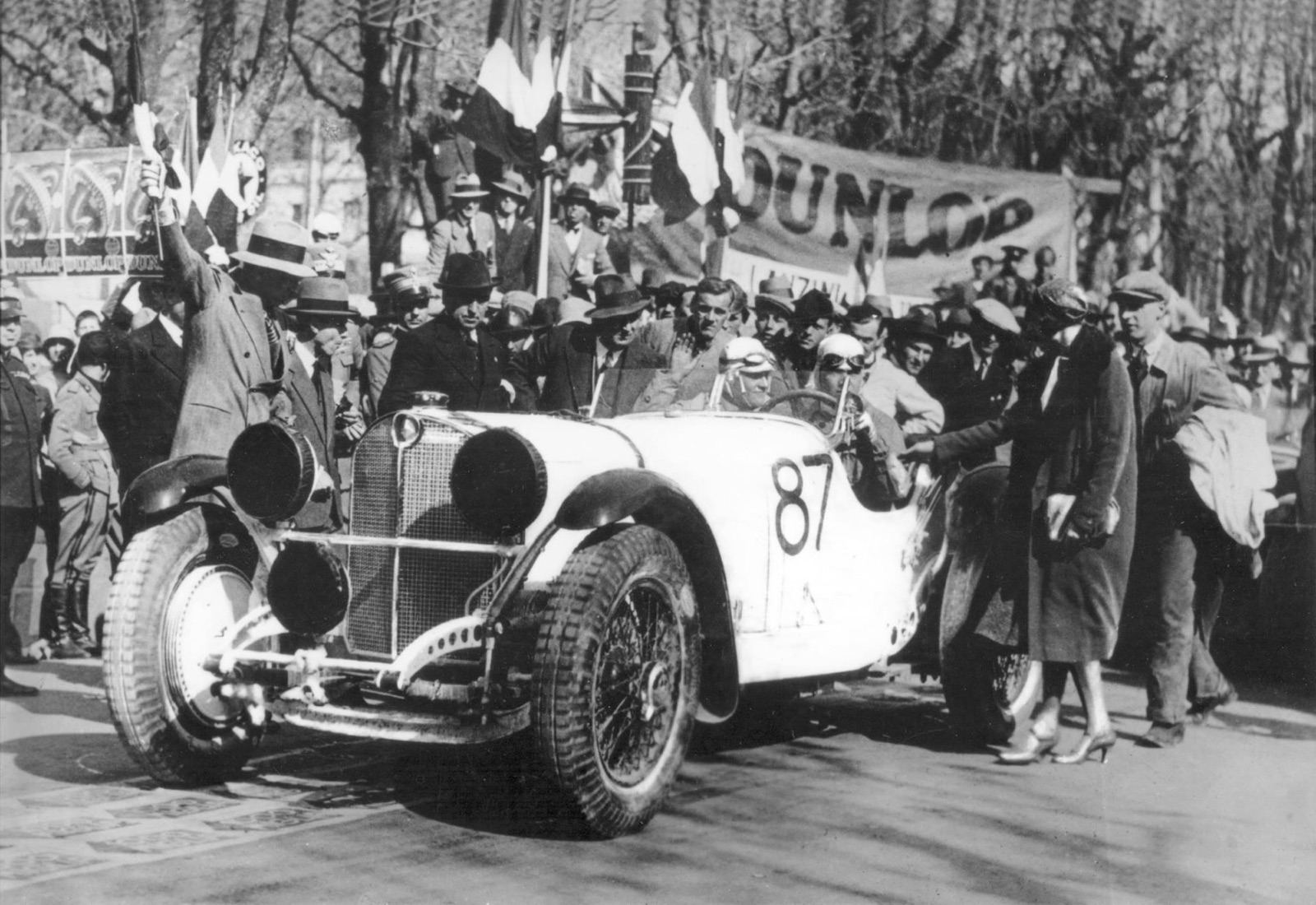 Two helmeted men in a white Mercedes-Benz numbered 87 surrounded by a crowd, a man waving a flag and a Dunlop banner behind, a woman in a long coat beside the car