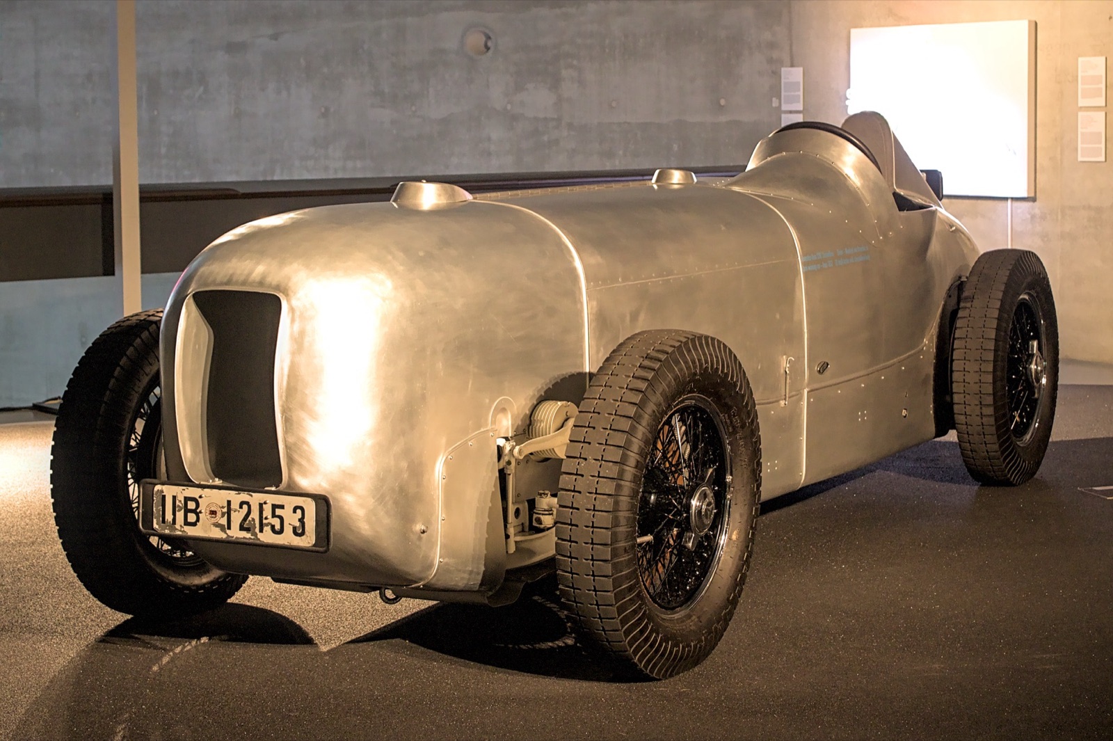 A long, bare-aluminium streamlined racing car with a rounded nose, an oval grille opening and a German number plate, on display indoors