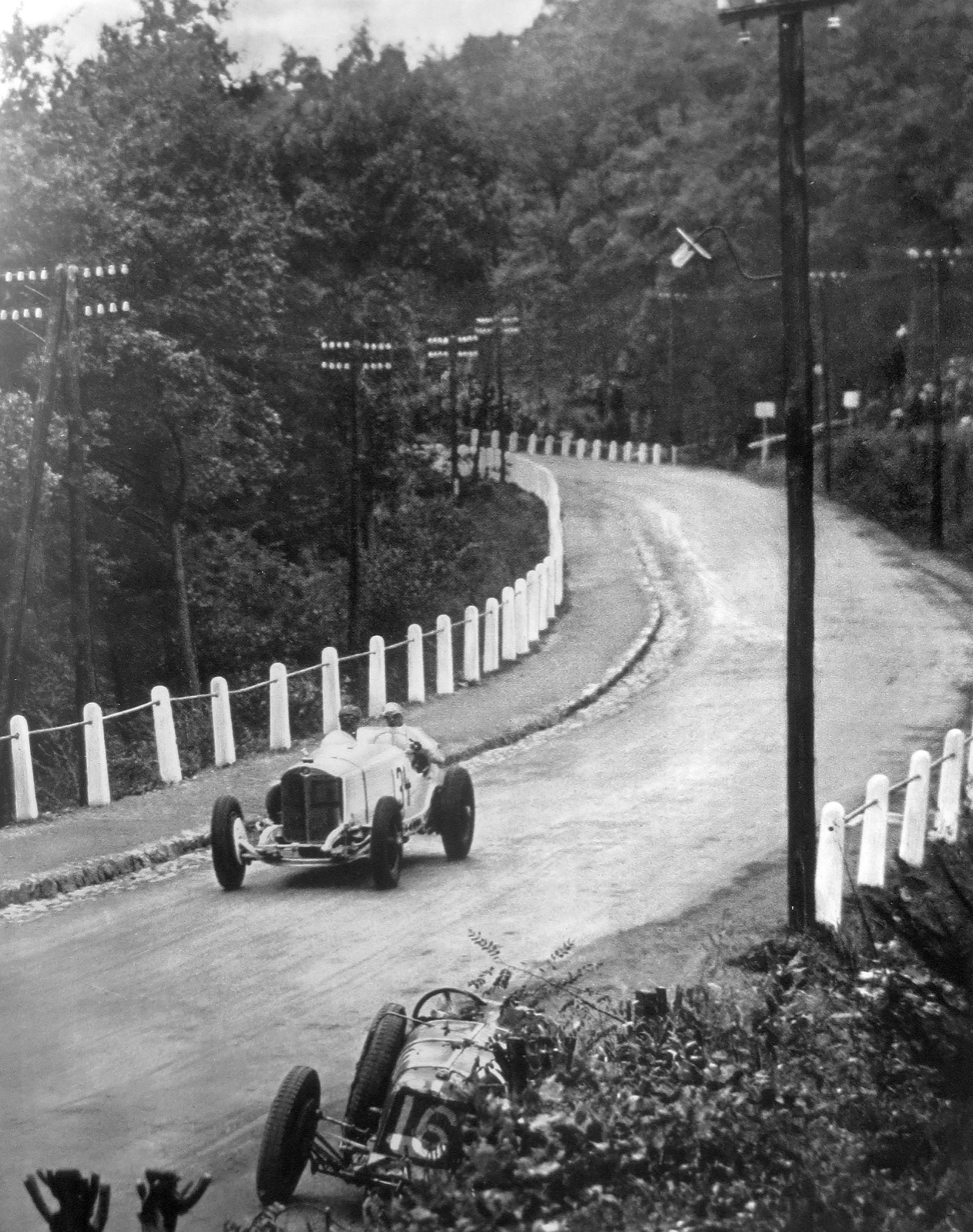 A white Mercedes-Benz with two men aboard rounding a bend on a tree-lined road with white posts, a second racing car at the roadside in the foreground