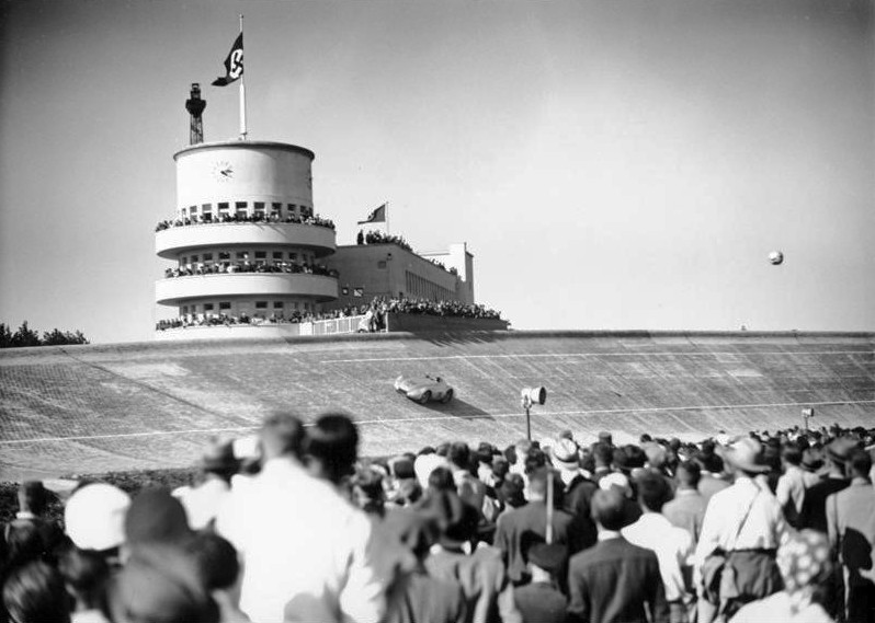 Crowds watch a streamlined racing car high on the steep brick banking of the Avus, below a round control tower flying flags