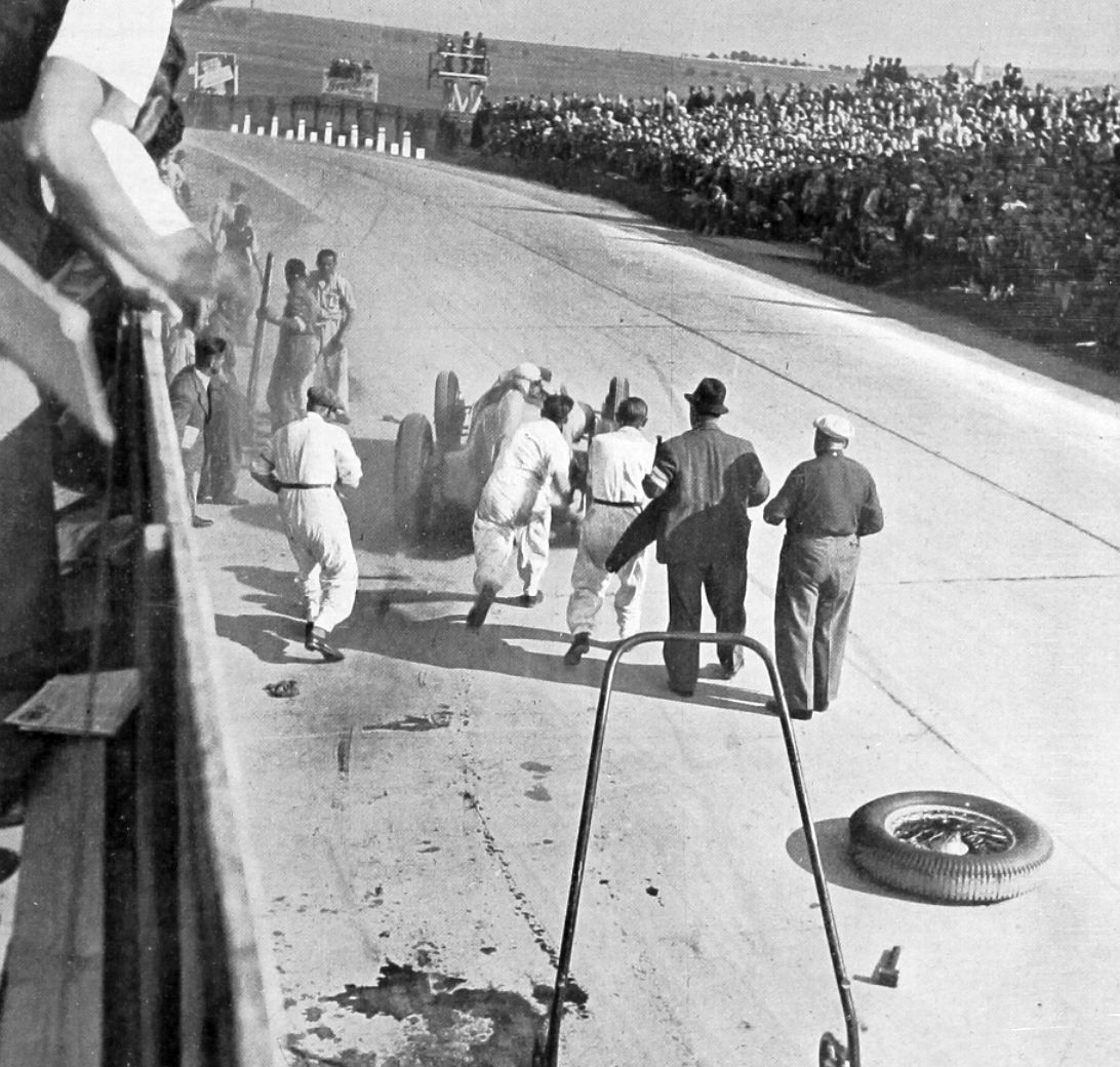 Mechanics in white overalls push Caracciola's Mercedes-Benz W125 away from the pits on a sunlit road circuit lined with spectators