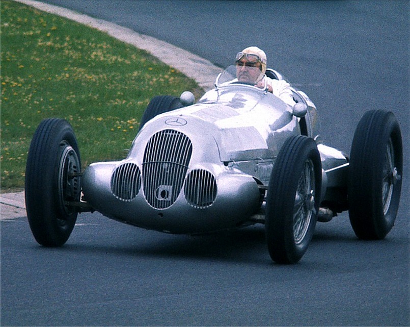 An elderly Hermann Lang in a cloth helmet and goggles drives a silver Mercedes-Benz W125 round a hairpin, showing its three nose openings