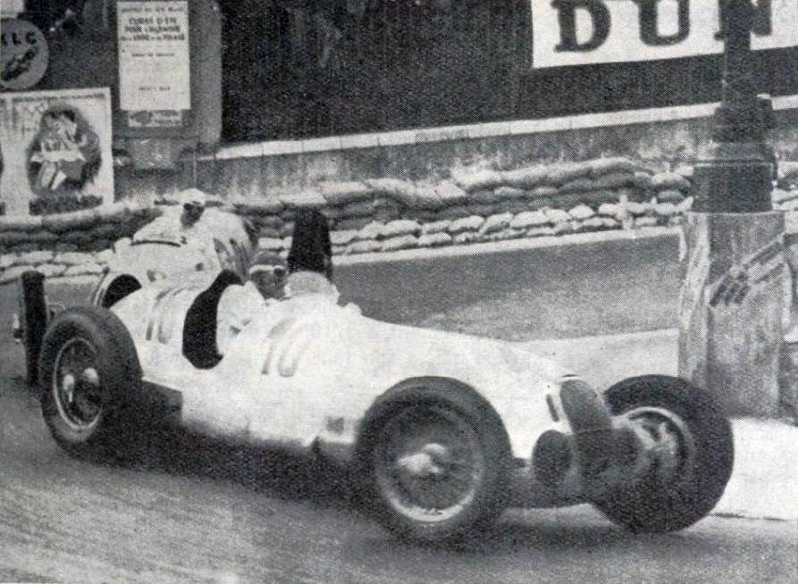 Two white Mercedes-Benz W125 Grand Prix cars cornering close together beside sandbags in the streets of Monaco