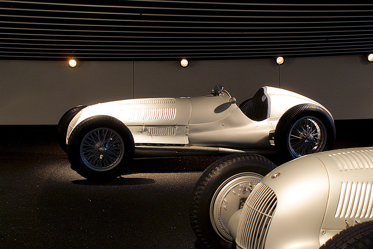 A silver Mercedes-Benz W125 Grand Prix car with wire wheels, lit in a dark museum hall, the nose of another racing car in the foreground