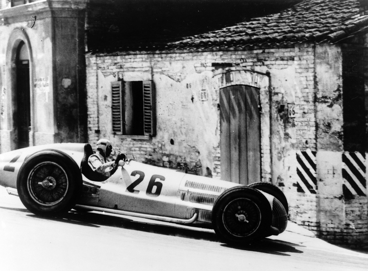 Rudolf Caracciola in a silver Mercedes-Benz W154 numbered 26 drives past an old brick house with a shuttered window