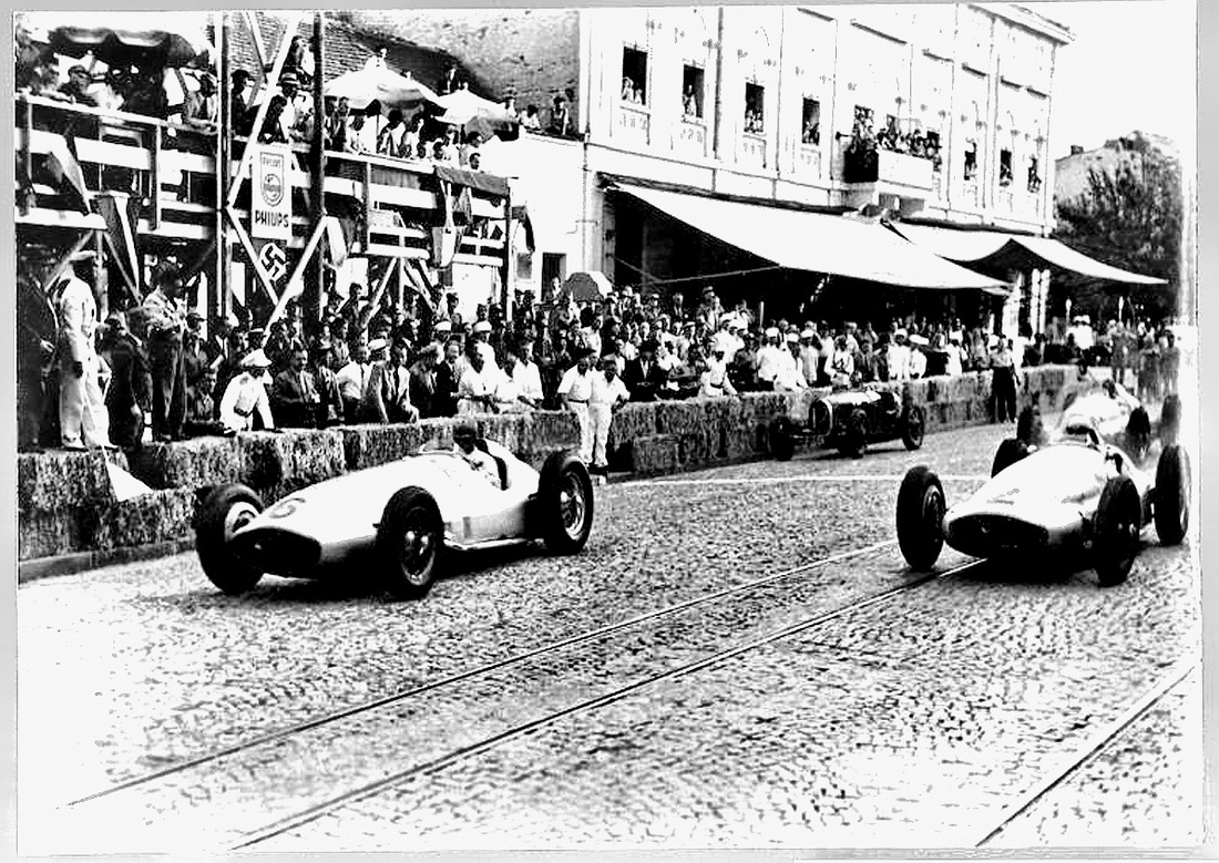 Two silver Mercedes-Benz W154 Grand Prix cars on a cobbled street with tram lines, watched by a crowd behind straw bales and from a wooden stand