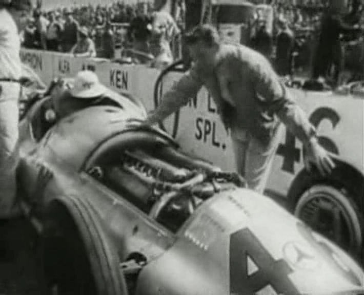 A man leans over the open engine bay of a silver Mercedes racing car with the three-pointed star on its nose, in front of a pit wall marked Don Lee Spl.