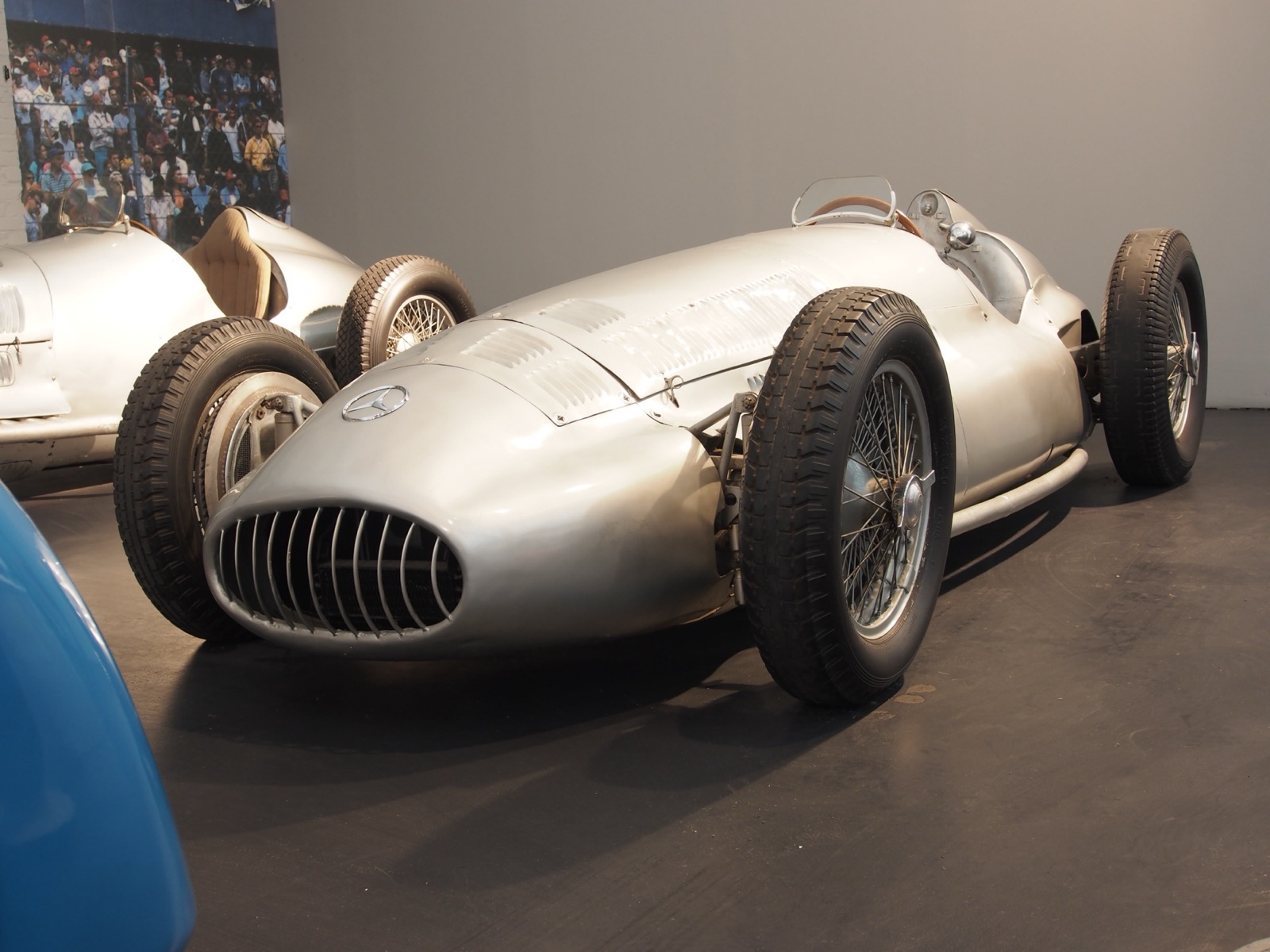 A silver 1939 Mercedes-Benz W154 with a rounded slatted nose and wire wheels on display in a museum hall