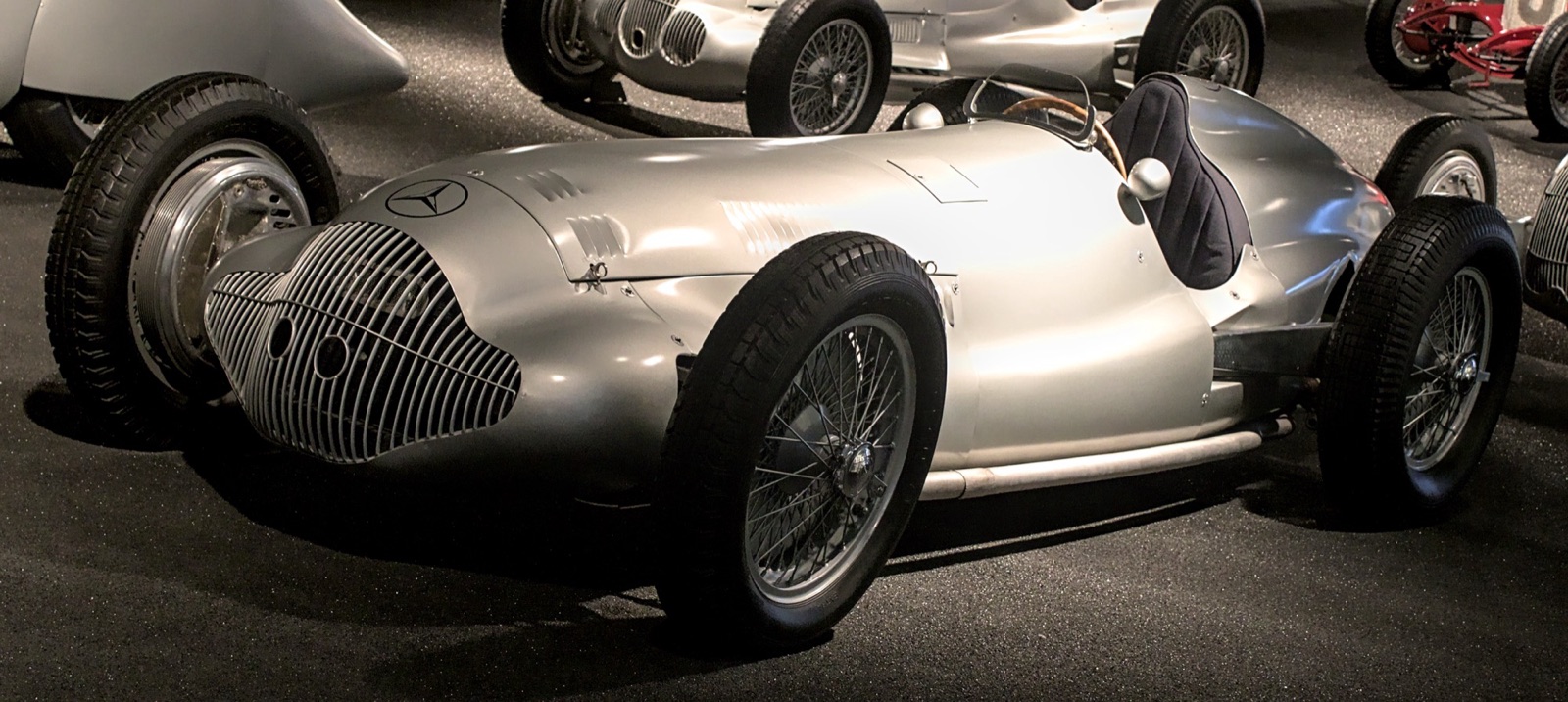 A silver Mercedes-Benz W154 Grand Prix car in its 1938 form, with a slatted grille and wire wheels, lit in a dark museum hall among other racing cars