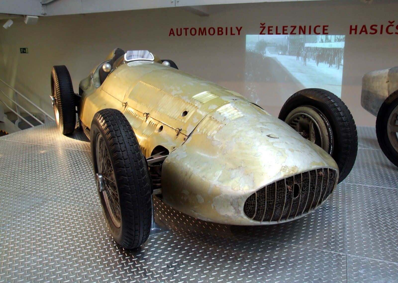 A Mercedes-Benz W154 with worn, patchy silver paint on a chequer-plate floor in a museum hall