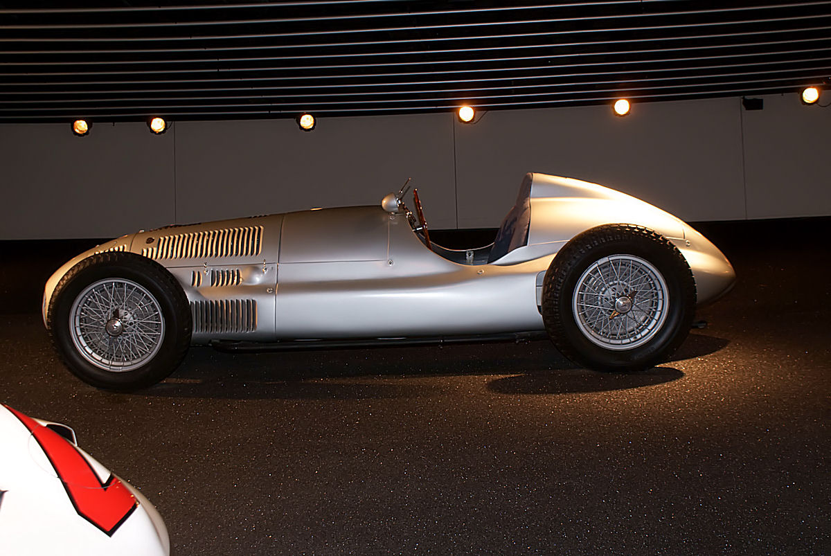 Left side view of a silver Mercedes-Benz W165 single-seater with wire wheels, louvred bonnet and short rounded tail, under spotlights