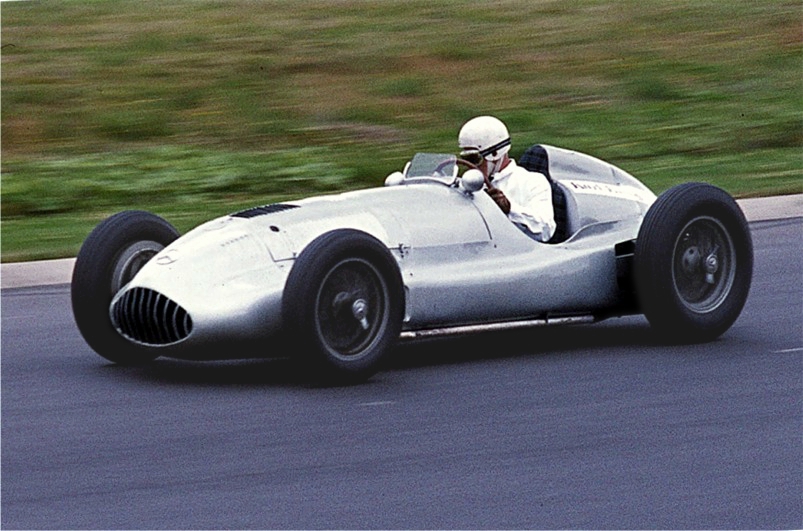 A silver Mercedes-Benz W165 with no race number, driven at speed on a track with a grass bank behind, the driver in a white helmet and overalls