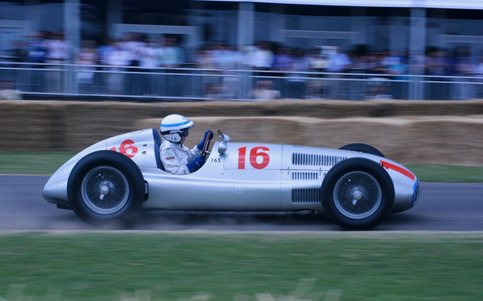 John Surtees in a white helmet driving a silver Mercedes-Benz W165 numbered 16 past straw bales and a grandstand
