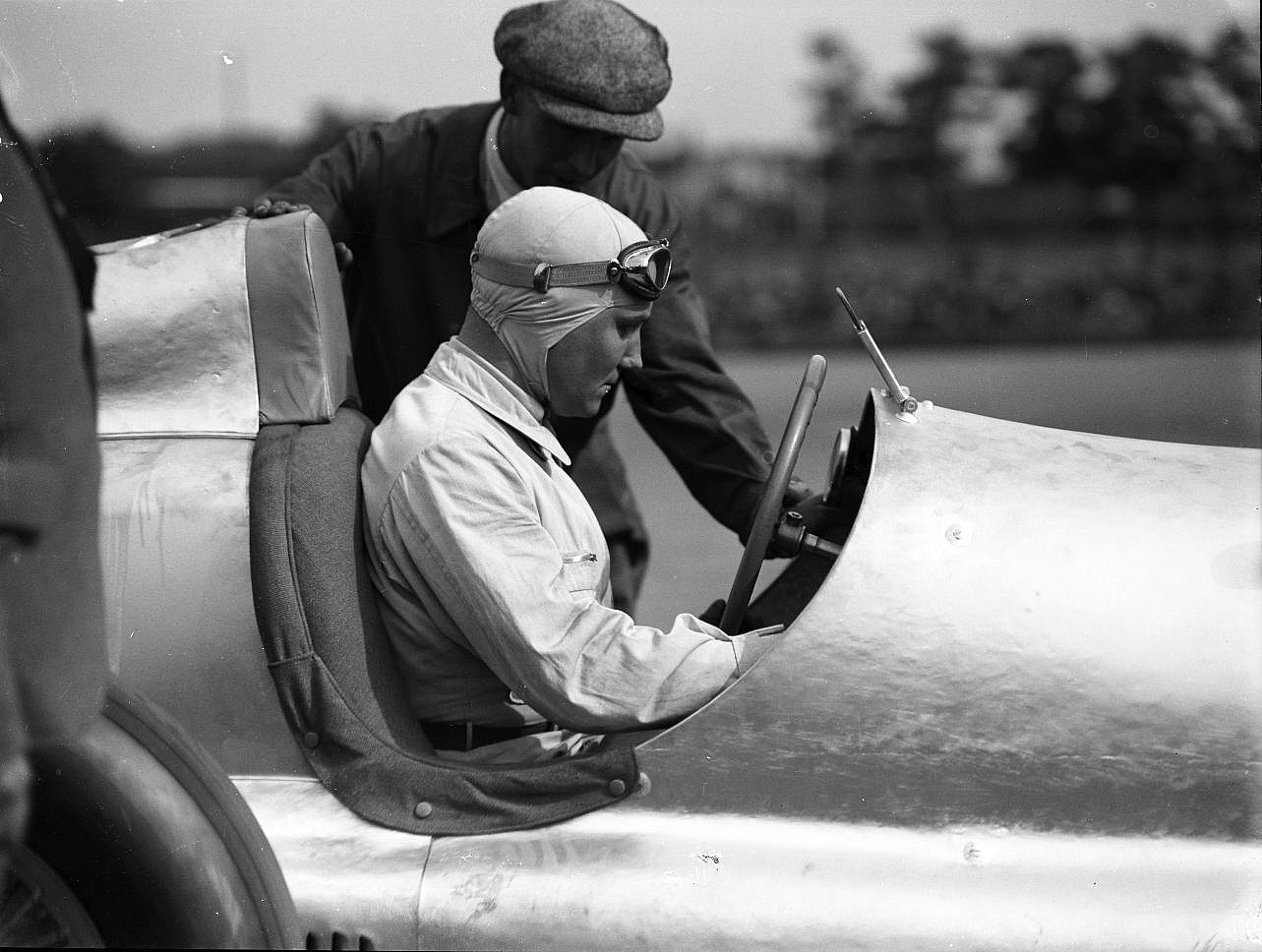 Manfred von Brauchitsch in a cloth helmet and goggles sits in the cockpit of a Mercedes-Benz W25 while a mechanic in a flat cap leans over the car