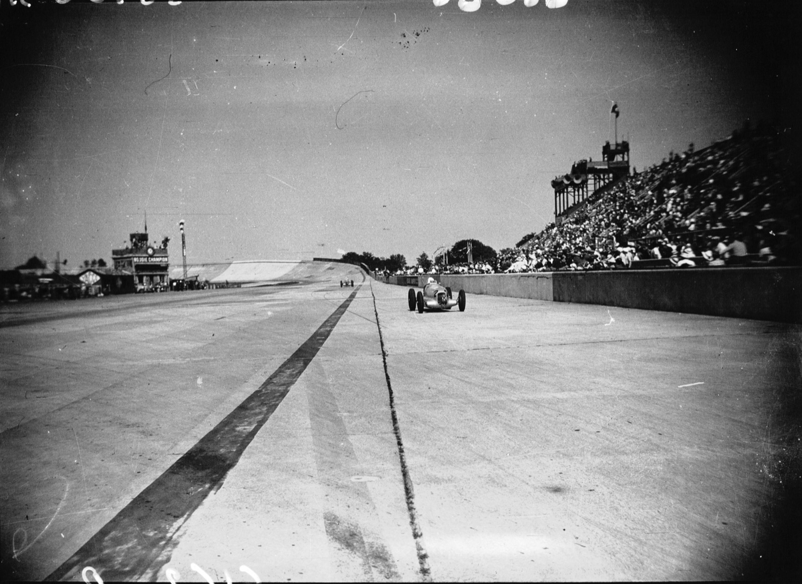 A Mercedes-Benz W25 on the wide concrete track at Montlhéry beside a packed grandstand, another car far behind