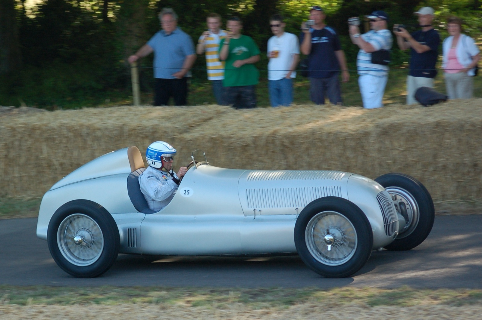 Jochen Mass in a white helmet drives a silver Mercedes-Benz W25 numbered 25 past straw bales and spectators with cameras