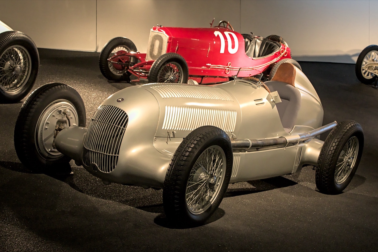 Silver Mercedes-Benz W25 Grand Prix car with wire wheels, a vertical-bar grille and louvred bonnet, displayed in a museum hall