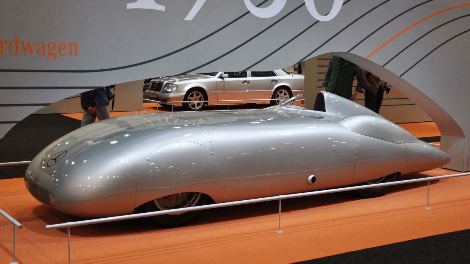 A long, low, fully enclosed silver Mercedes-Benz record car with covered wheels and a small cockpit fairing, on an exhibition stand