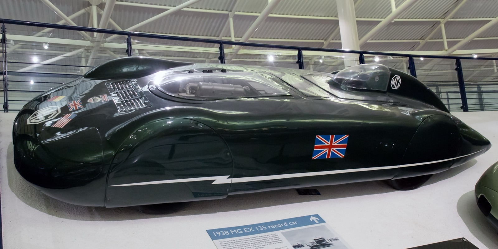 The dark green, fully enclosed MG EX135 record car on display at Gaydon, with a Union flag on its side and a display board in front