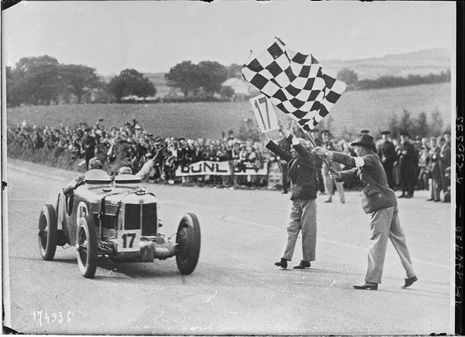 Nuvolari's MG K3 Magnette, number 17, taking the chequered flag on the Ards circuit, with two officials waving flags and a crowd behind a Dunlop banner