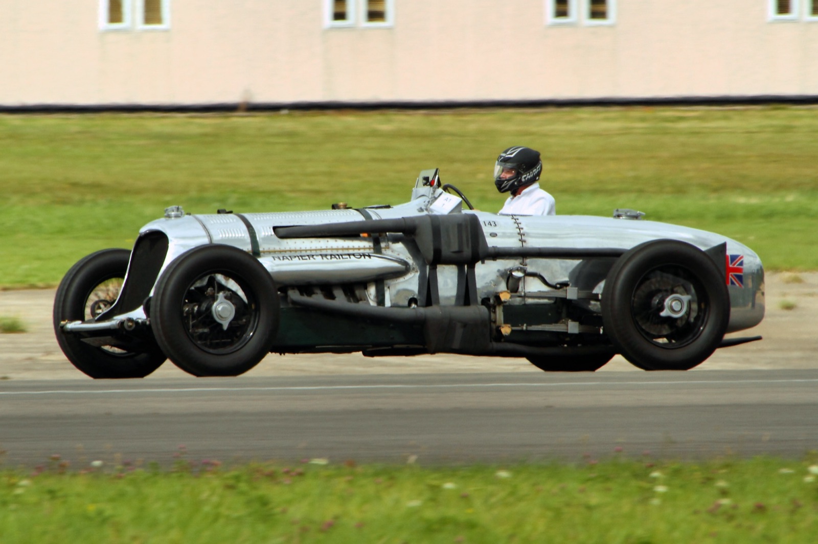 The silver Napier-Railton running along the runway at Dunsfold, its driver in a black helmet, with grass and a hangar wall behind