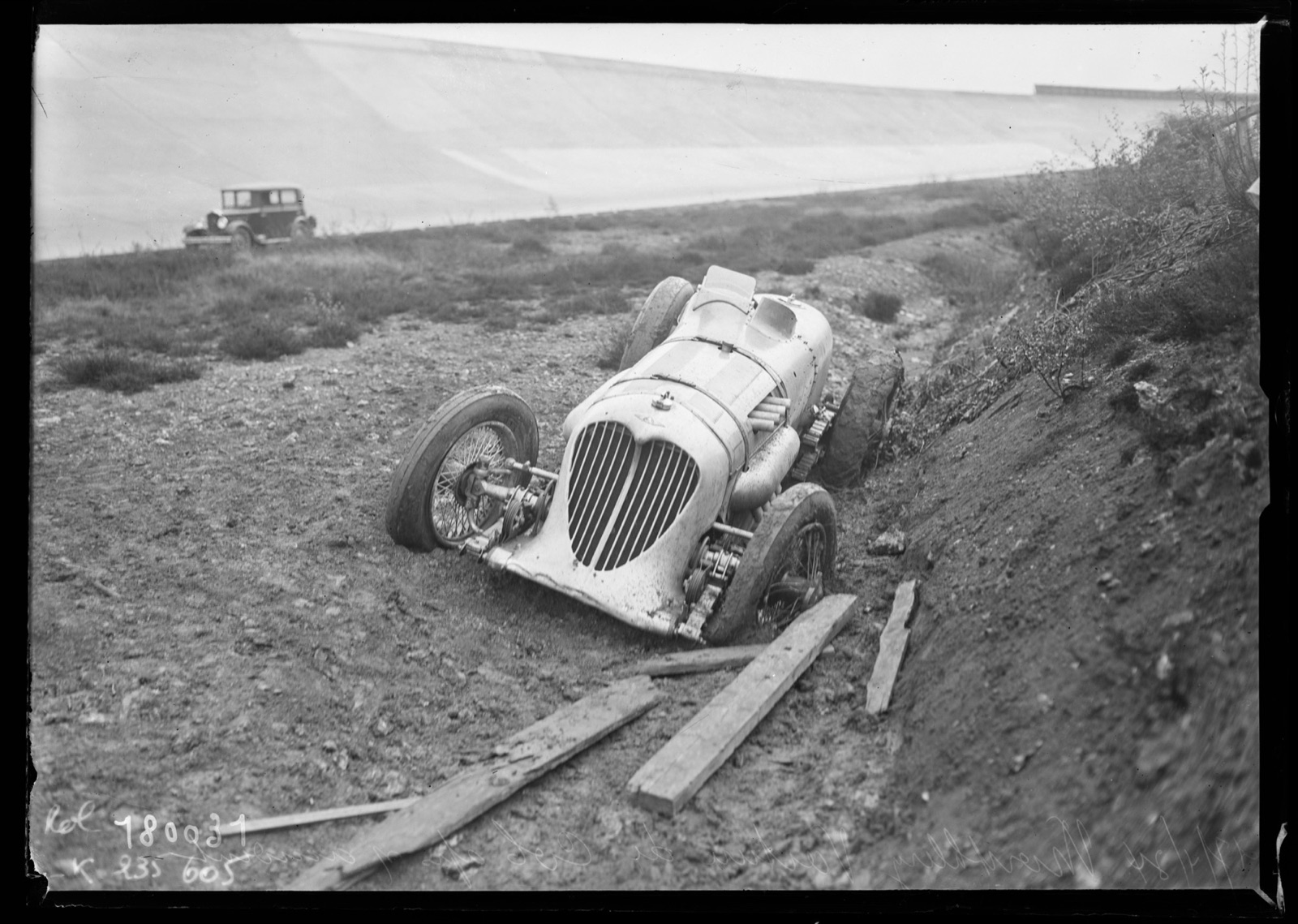 Black-and-white press photograph of the Napier-Railton lying nose-up in a ditch below the Montlhéry banking, with a saloon car on the track behind