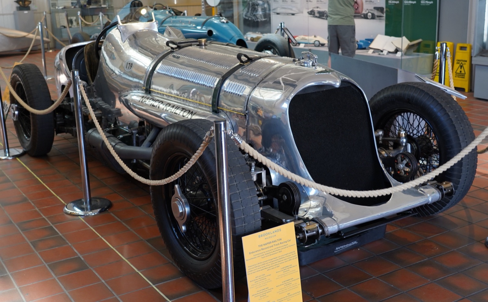 The polished Napier-Railton on display behind rope barriers at Brooklands Museum, with an information board in front